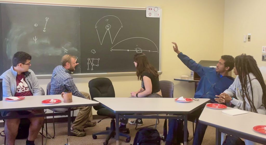 Several students and a faculty member sit at tables. They are turning toward the chalkboard where diagrams have been drawn, and discussing the ideas on the board.