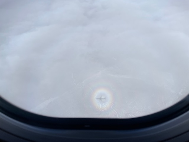 A full circle rainbow of colors surrounds the shadow of a plane on clouds.