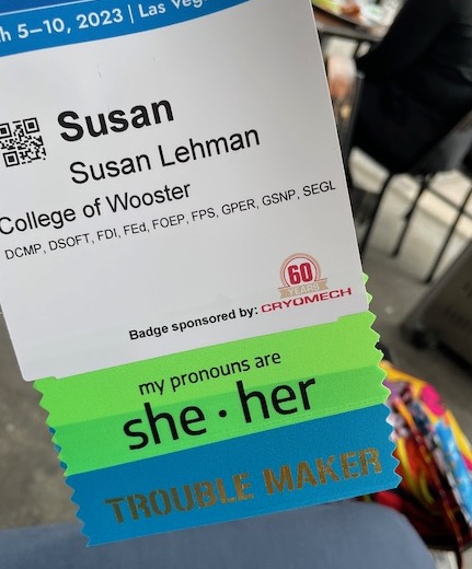 Name tag with ribbons that say "she her" and "Trouble Maker"