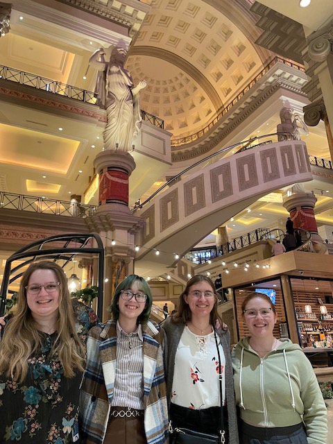Four women stand together in a grand atrium space designed to look Roman.