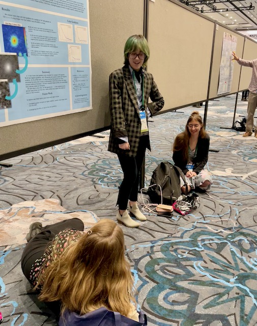 A woman stands and looks at two women sitting on the ground at the poster session.