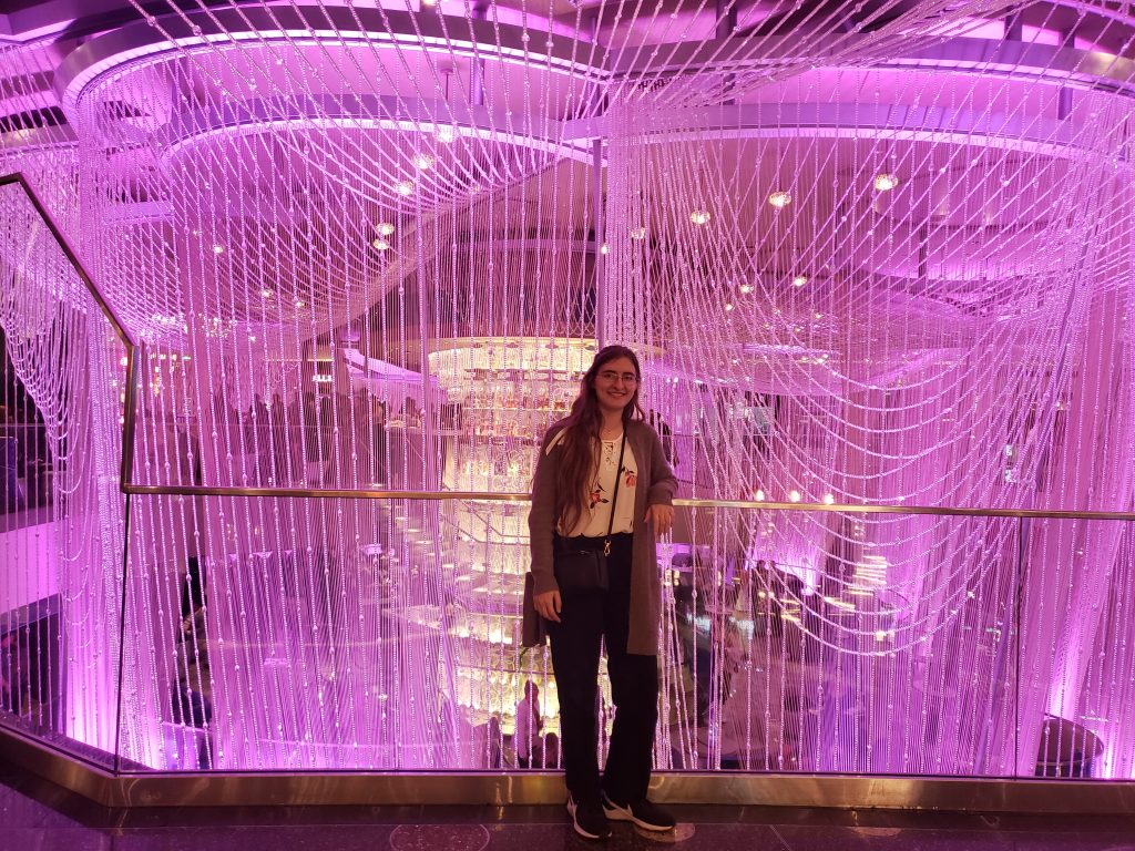 A woman stands in front of a large pink chandelier.