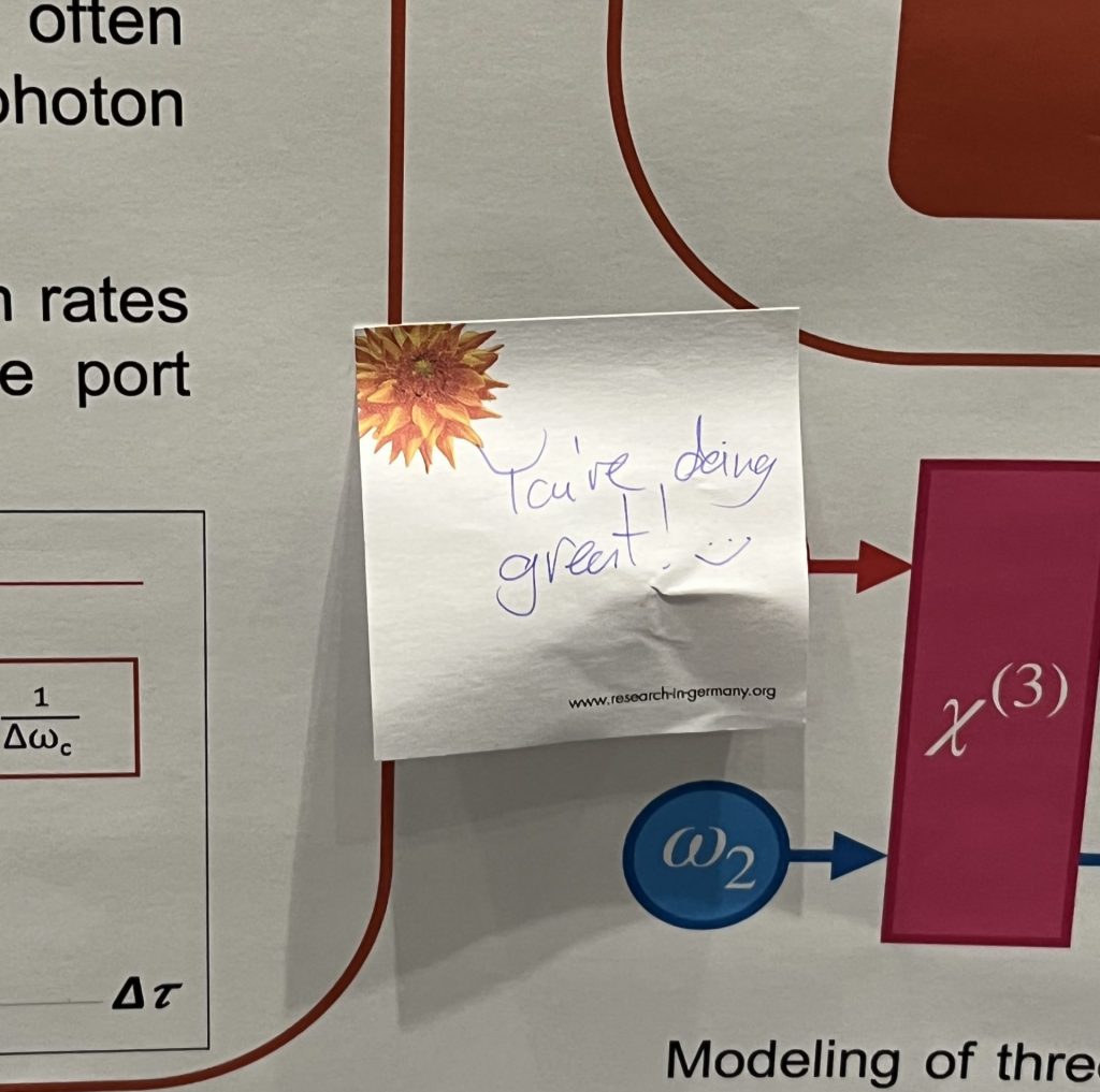 A sticky note reading "You're doing great" with a smiley face is stuck to a scientific poster.